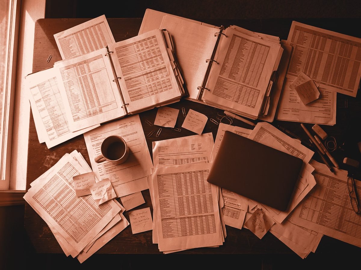 Bureau encombré de feuilles de calcul imprimées, de classeurs ouverts et de notes autocollantes, vu du dessus